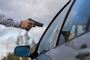 mão masculina apontando arma de fogo para vidro de carro blindado
