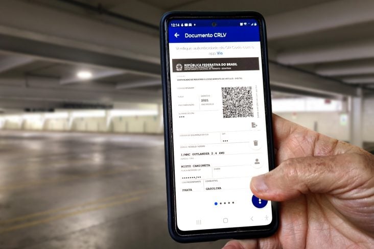 mao masculina segurando celular com o documento do carro digital ou crlv-e aberto em estacionamento fechado CRLV 2024