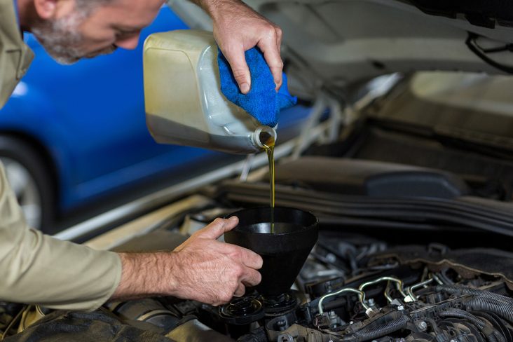 Homem branco segurando com a mão esquerda um galão de óleo e colocando no motor com um funil. Primeira troca de óleo