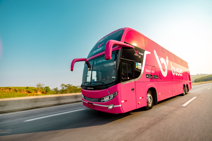 Ônibus rosa da Buser em estrada com ceú azul ao fundo visto da diagonal mostrando frente e lateral do veículo