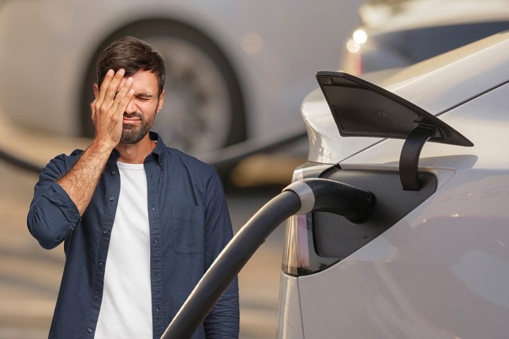 Homem com a mão no rosto de arrependimento ao lado do carro elétrico recarregando