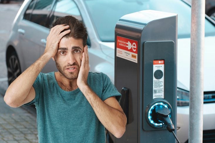 Homem com as mão no rosto com a expressão de cansado. Ao fundo um carro elétrico recarregando.