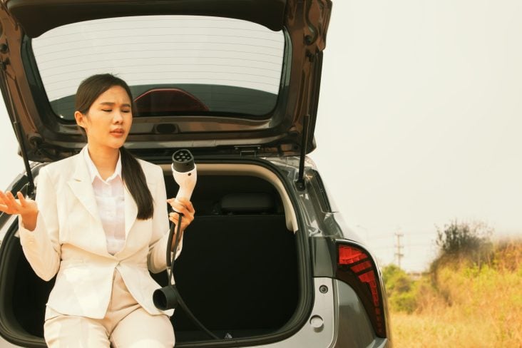 Shutterstock seguro de carro elétrico é mais caro Mulher asiática vestida de branco sentada no porta malas aberto do carro segurando um carrgador de veículo elétrico