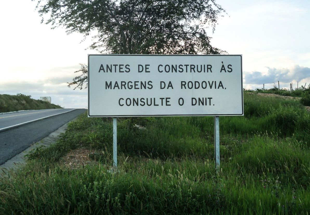 Placa de trânsito educativa com fundo branco – “antes de construir às margens da rodovia consulte o DNIT”