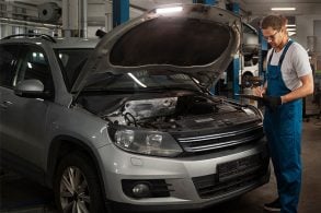 Carro com capô aberta na oficina mecânica; mecânico anota em uma prancheta