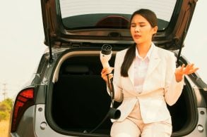 Mulher asiática vestida de branco sentada no porta malas aberto do carro segurando um carrgador de veículo elétrico