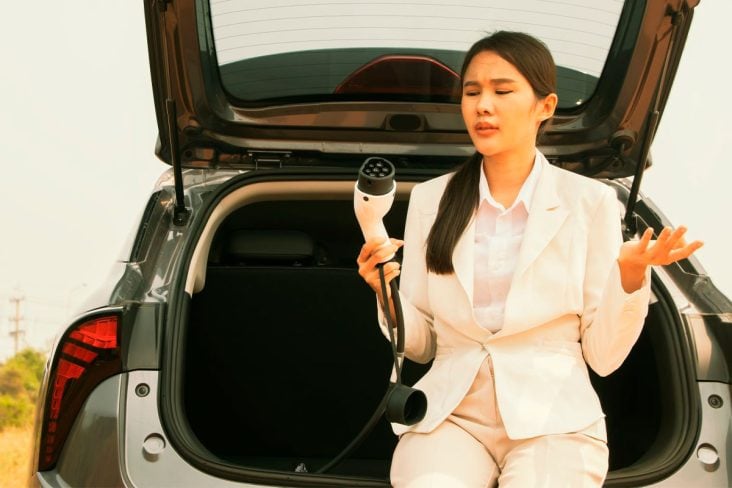 Mulher asiática vestida de branco sentada no porta malas aberto do carro segurando um carrgador de veículo elétrico