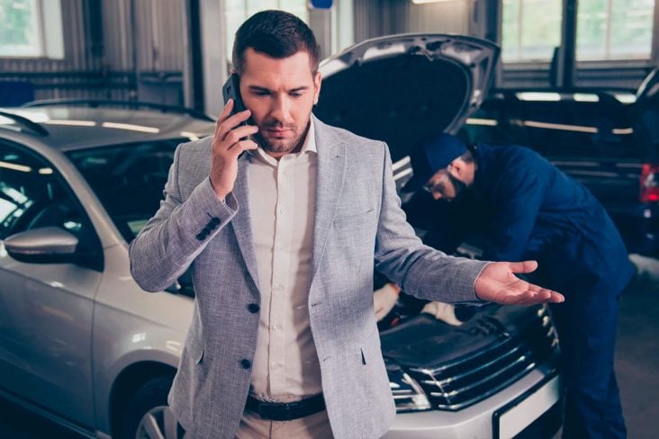 Homem ao telefone com expressão de indignação e ao fundo mecânico consertando o motor