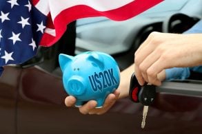 Homem dentro de um carro segura na mão direita um cofrinho azul e na mão esquerda a chave do carro, bandeira americana na lateral da imagem