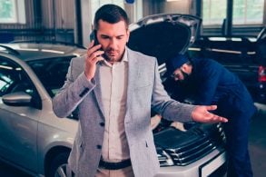 Homem ao telefone com expressão de indignação e ao fundo mecânico consertando o motor