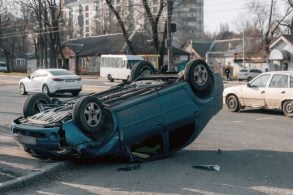 shutterstock capotamento carro cinza escuro capotado em rua com carros estacionados e casas ao fundo